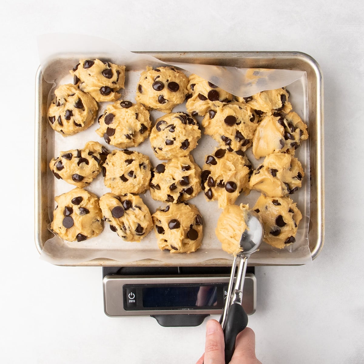Scoops of chocolate chip cookie dough on a parchment-lined baking sheet set on a kitchen scale with a hand holding a spring loaded ice cream scoop transferring a portion of dough onto the baking sheet.