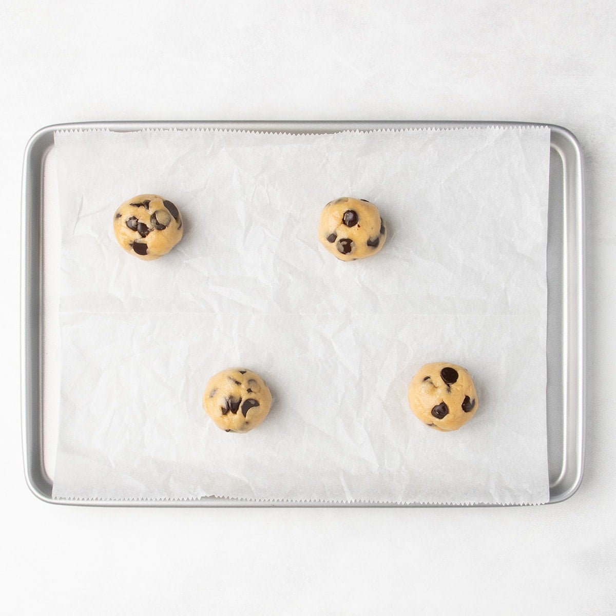 Four smooth balls of chocolate chip cookie dough spaced on a parchment-lined baking sheet.