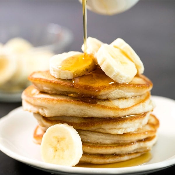 closeup of maple syrup pouring from a small white pitcher over a tall stack of golden brown pancakes with banana slices