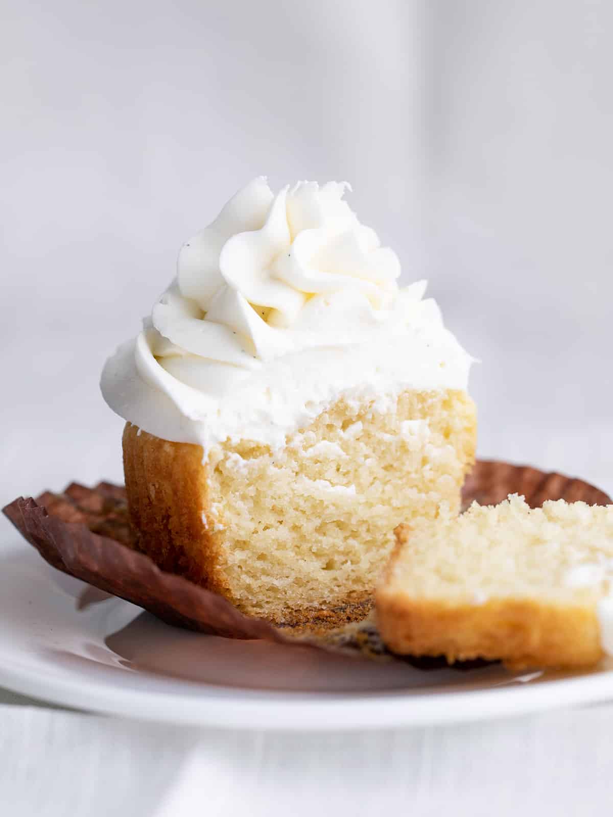 One gluten free vanilla cupcake with swirled white frosting cut in two pieces on brown paper liner on a small white plate above a white cloth