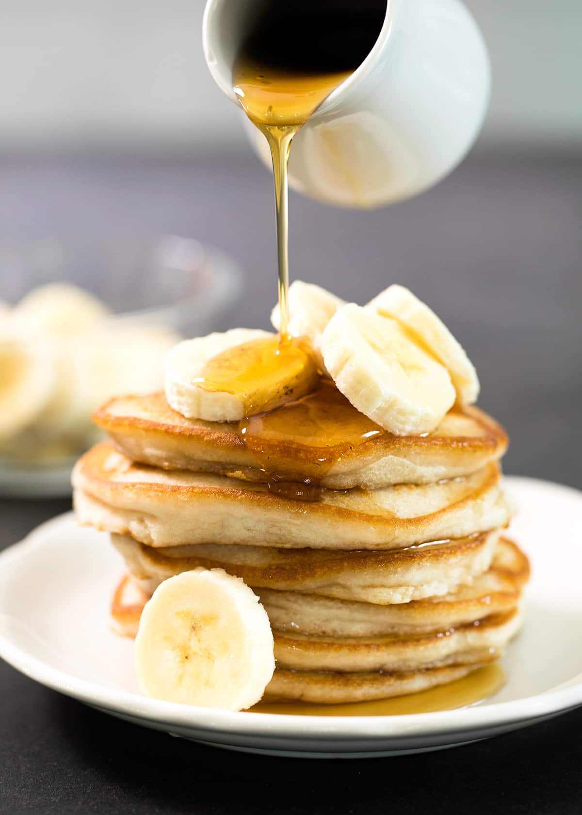 syrup pouring out of small white creamer onto stack of 6 syrupy golden brown pancakes with banana slices on white plate on black surface