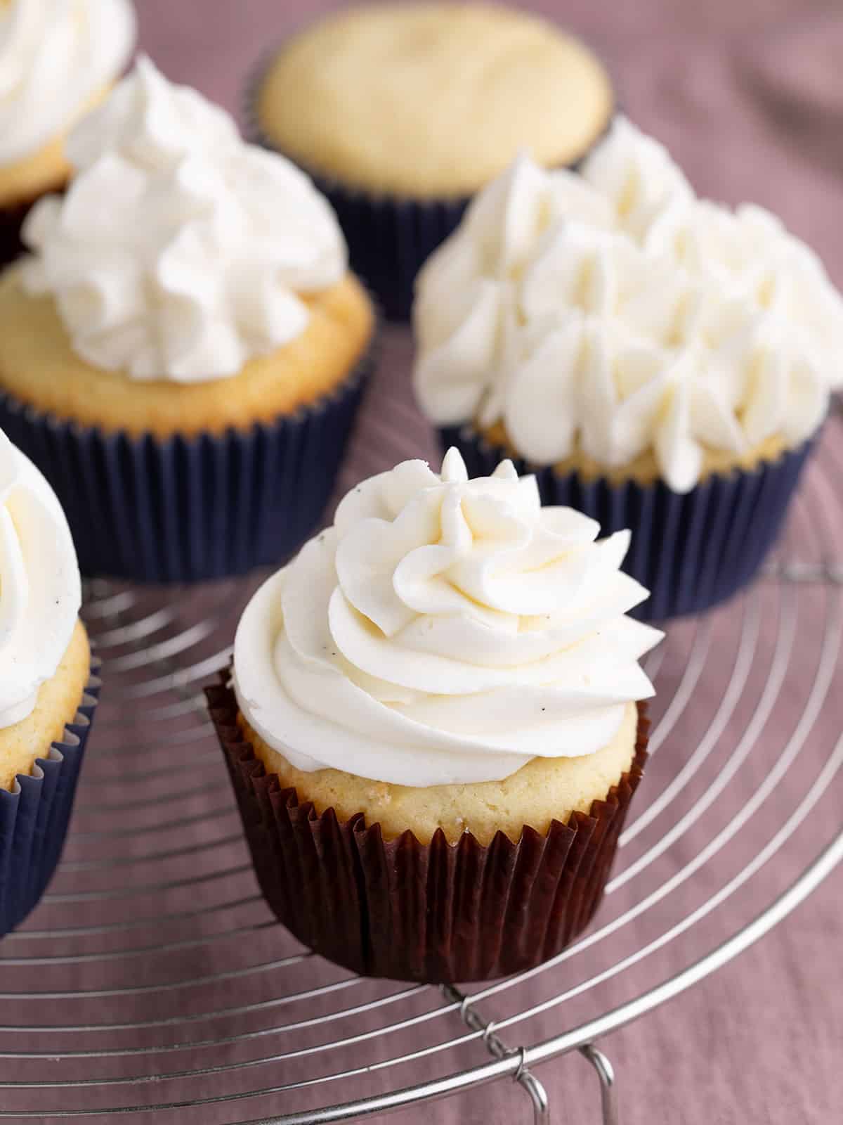 Yellow cupcakes with frosting in paper liners on round wire rack above purple cloth