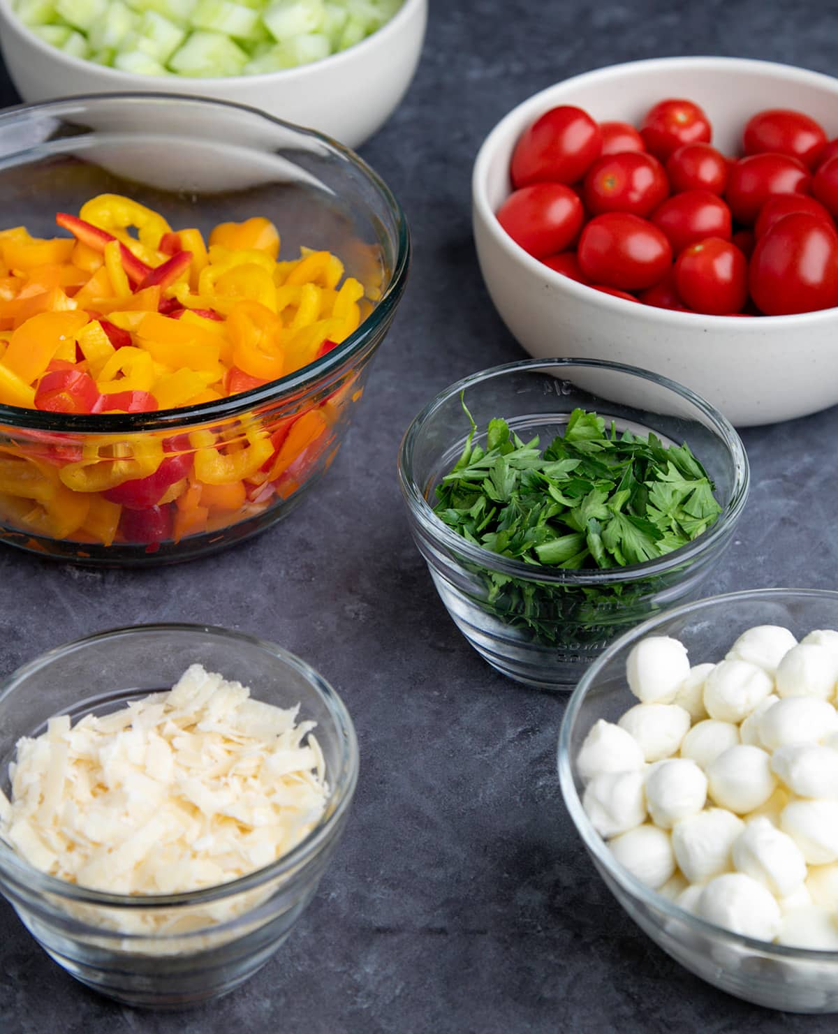 Bowls of chopped red and orange sweet pepper, cherry tomatoes, chopped scallions, parsley, and white small pearls of cheese on gray surface