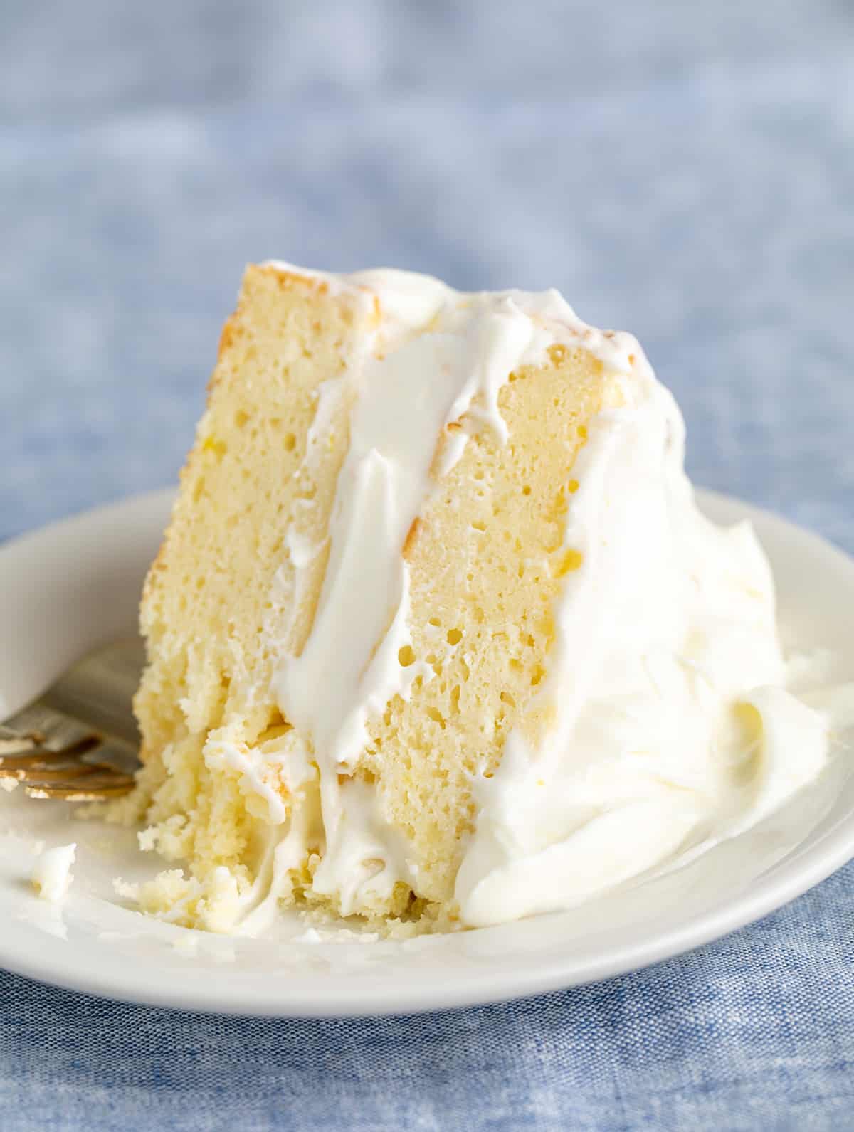 fork on side of slice of 2 layer lemon cake with lots of white frosting on small white plate on blue cloth