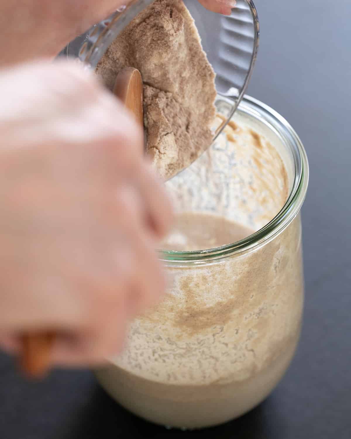 hands adding light brown flour to glass jar of light brown gluten free sourdough starter