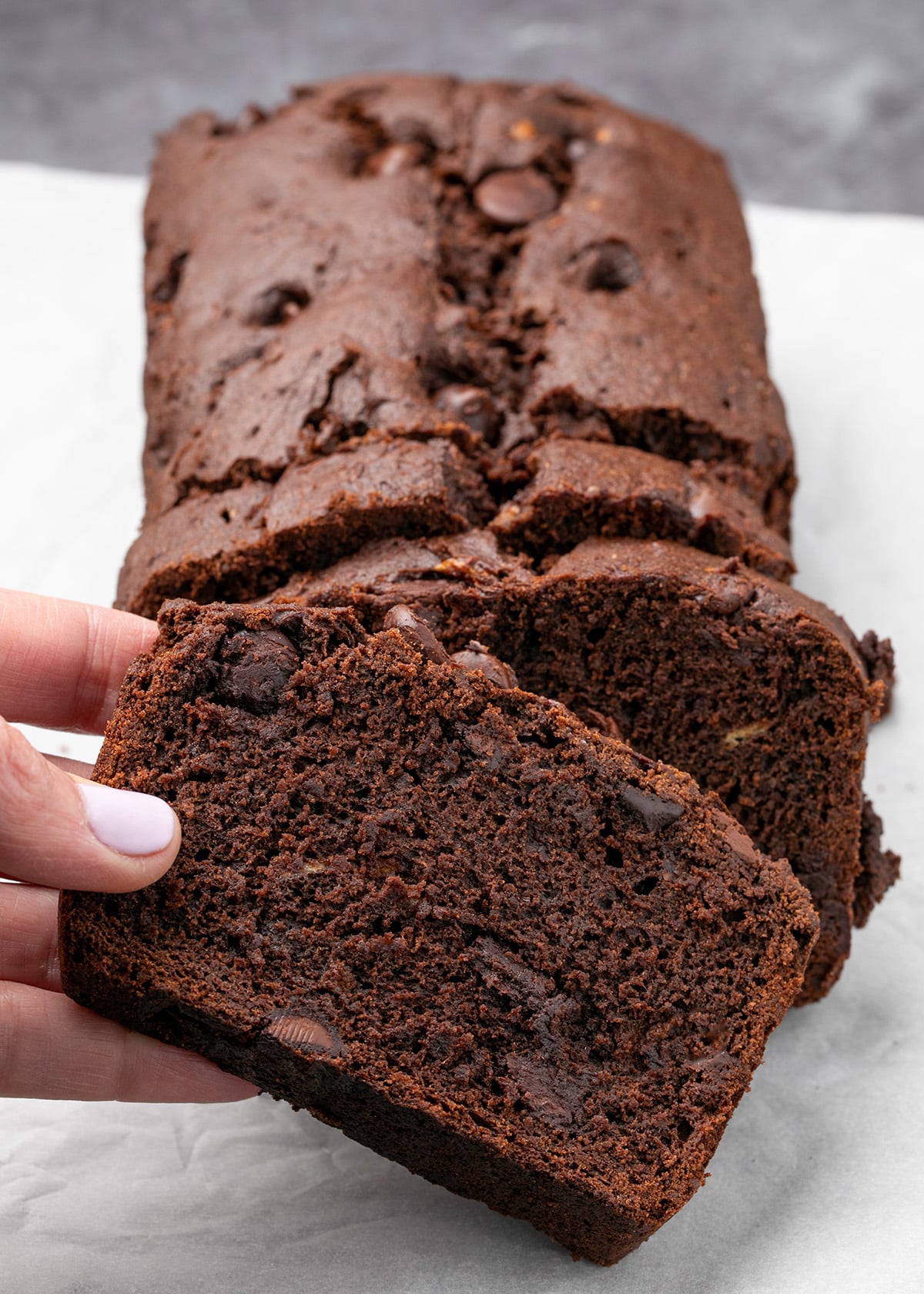 Fingers picking up slice of brown quick bread with chocolate chips from rest of loaf on white paper
