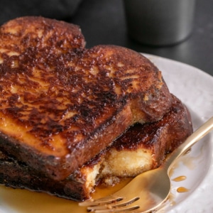 closeup image of stack of 2 slices of gluten free French toast with maple syrup on a white plate with one bite taken from one and a gold fork