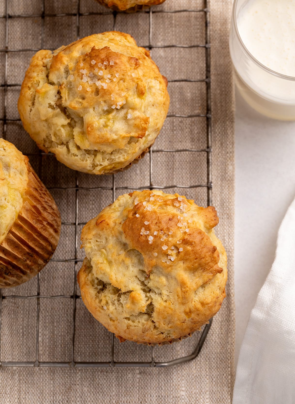 Two golden-brown muffins with coarse sugar on top rest on a wire cooling rack beside a glass of milk and a white cloth.