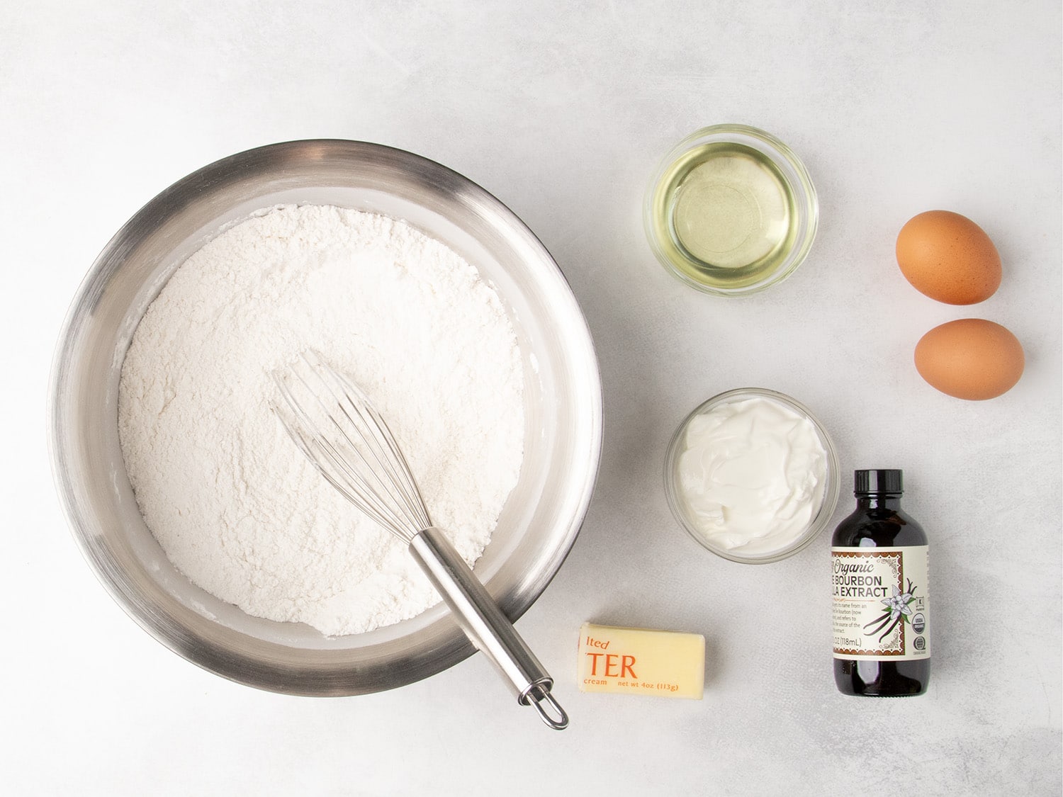 A whisk rests in a stainless steel bowl filled with white dry ingredients, beside bowls of oil and sour cream, two eggs, butter, and a jar of vanilla.