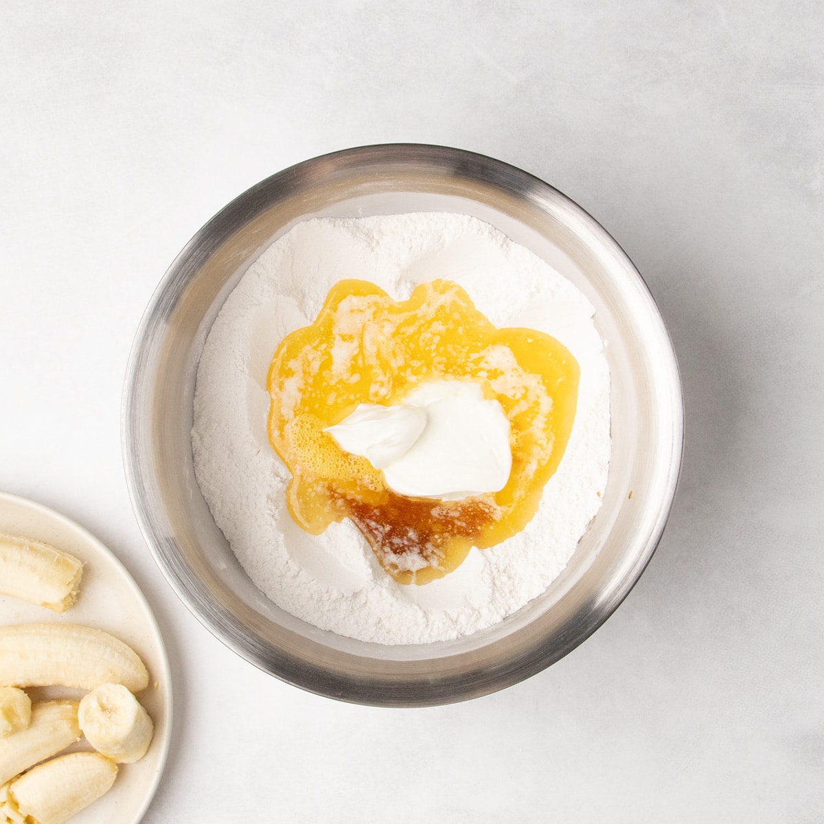 Melted yellow liquid, white sour cream, and brown liquid sit in the center of white dry ingredients in a stainless steel bowl, with peeled bananas on a plate nearby.