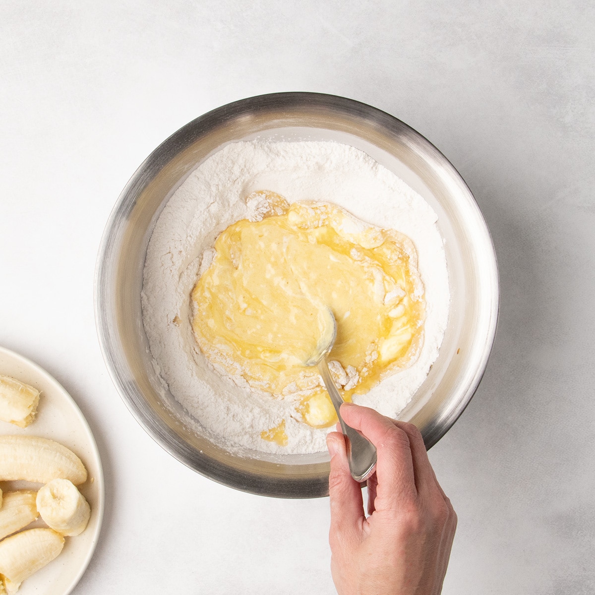 A hand uses a spoon to stir the wet ingredients together first before incorporating them into the dry ingredients in a stainless steel bowl.