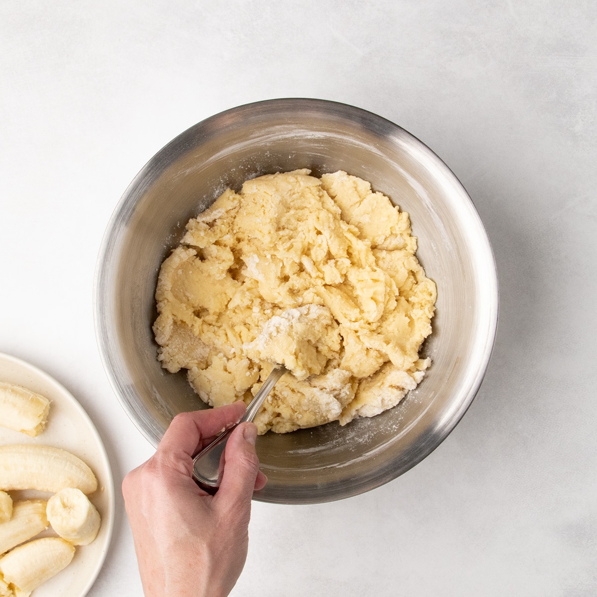 A hand stirs a thick, pale yellow mixture in a stainless steel bowl.