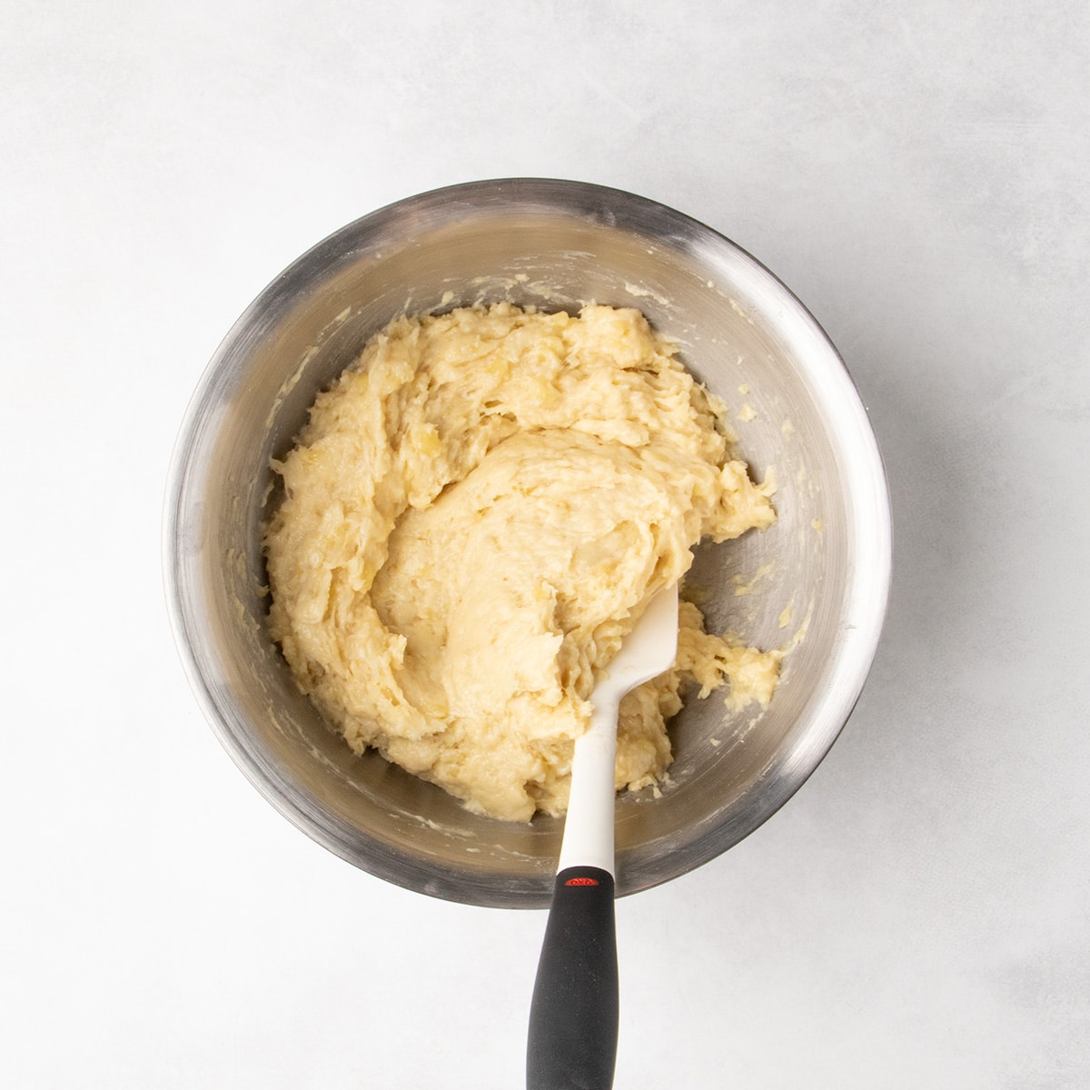 A white spatula rests in thick, pale yellow batter with visible mashed banana pieces in a stainless steel bowl.
