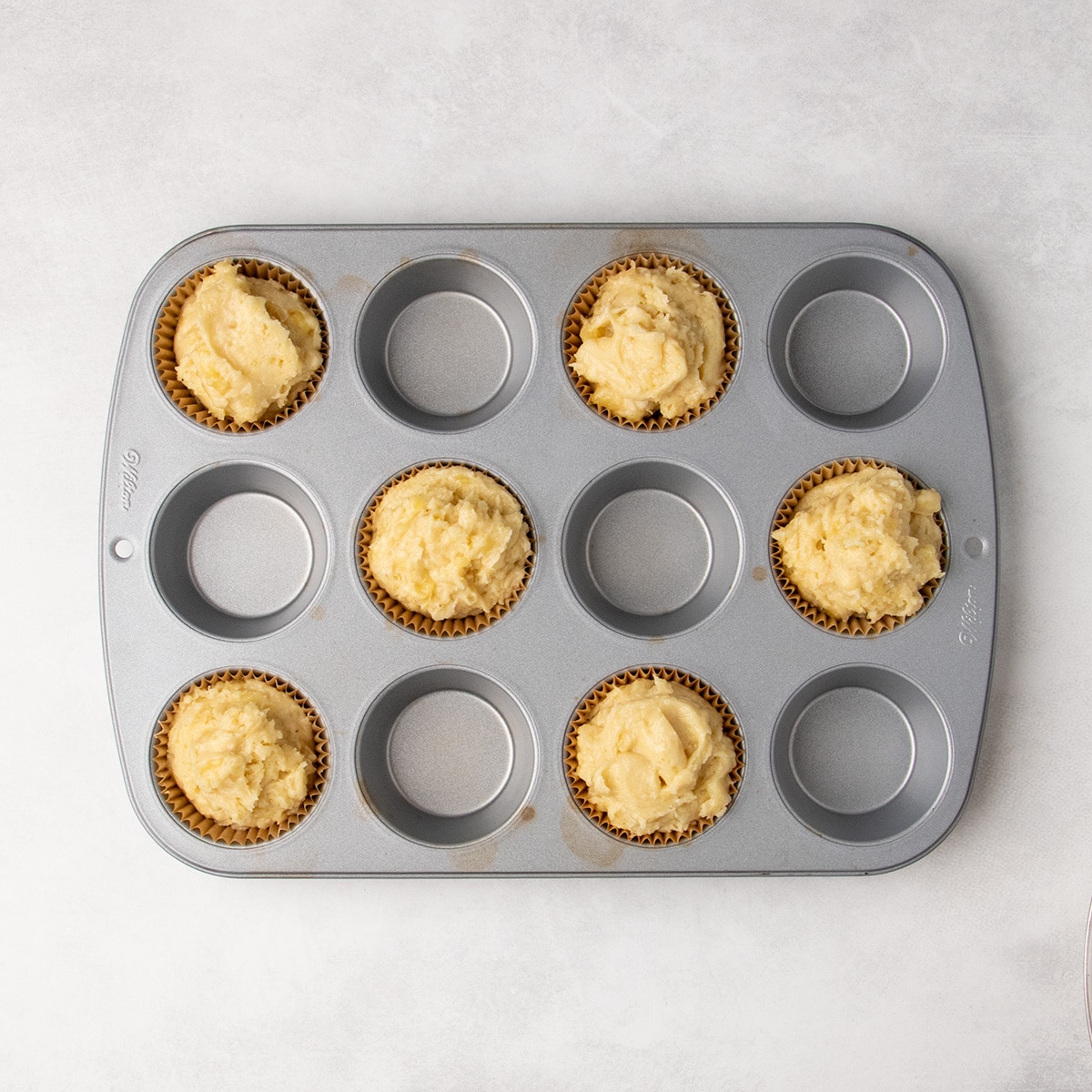 Six paper liners filled with pale yellow batter sit in alternating cups of a metal muffin pan lined with brown paper liners.