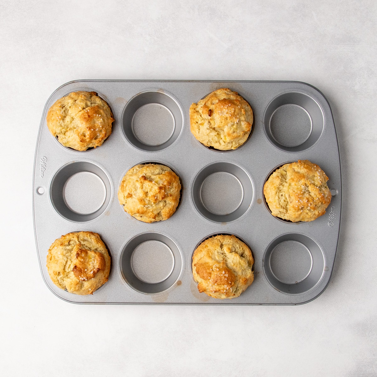 Six golden-brown banana muffins with rounded tops sit in alternating cups of a metal muffin pan.