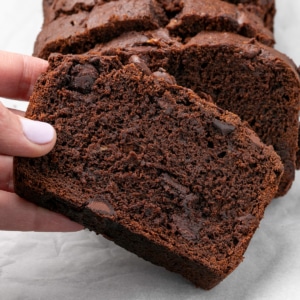 fingers picking up a slice of dark brown gluten free chocolate banana bread on white paper