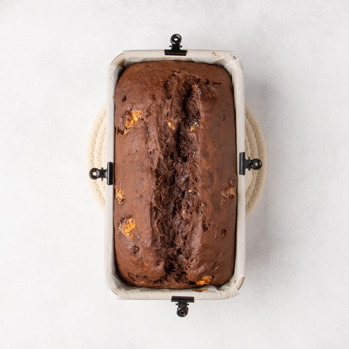 A baked chocolate loaf with a split top and visible banana pieces sits in a parchment-lined loaf pan with black clips on all 4 sides of the pan, securing white parchment paper.