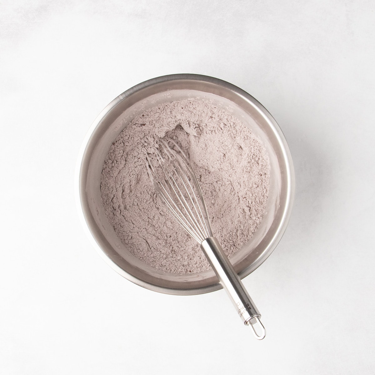 A whisk rests in pale brown dry ingredients combined in a stainless steel mixing bowl.