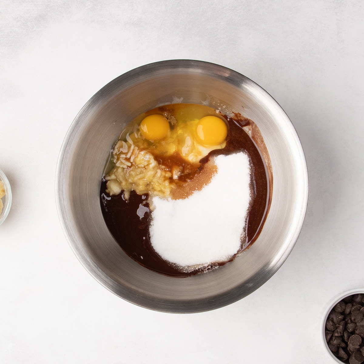 Two raw eggs, mashed banana, white sugar, and melted butter and chocolate sit together in a stainless steel mixing bowl.