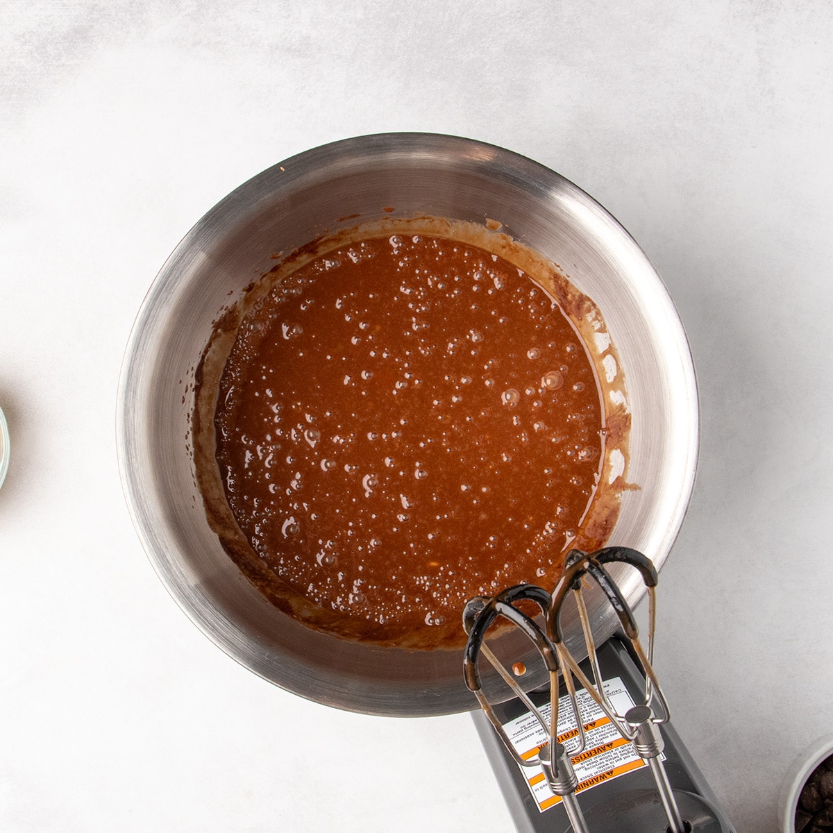 An electric hand mixer rests beside raw brown batter with tiny bubbles in a stainless steel mixing bowl.