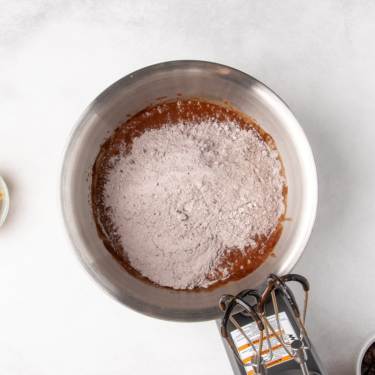 Pale brown dry ingredients cover part of the raw liquid brown batter in a stainless steel mixing bowl.