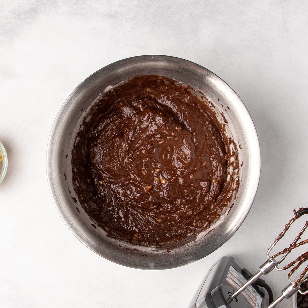 Thick brown chocolate batter with some pieces of mashed banana visible fills a stainless steel mixing bowl beside the beaters of an electric hand mixer.