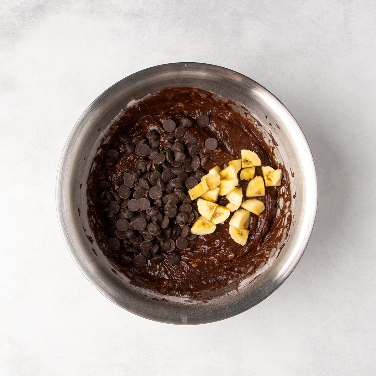 Chocolate chips and chopped banana sit on top of thick chocolate batter in a stainless steel mixing bowl.