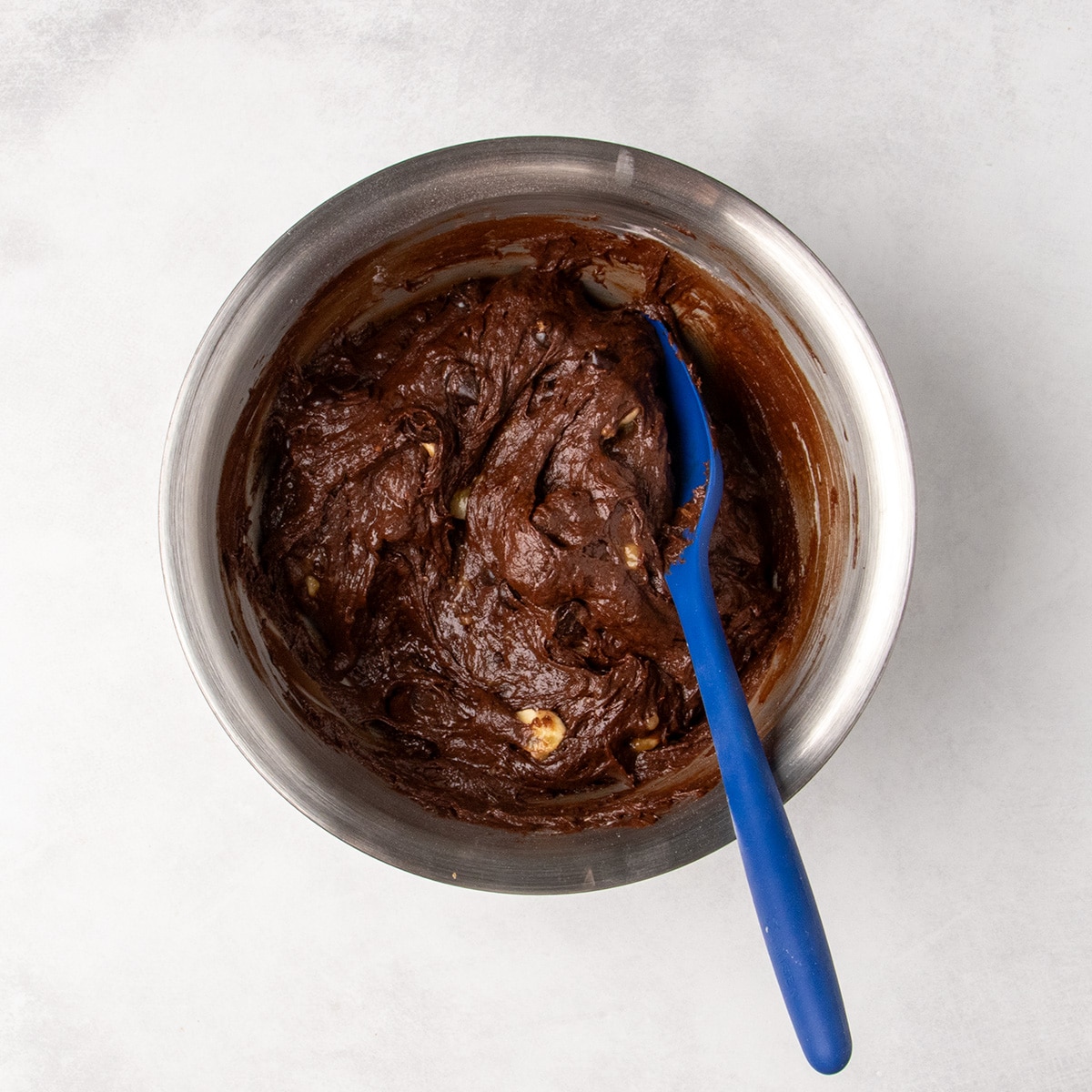 A blue silicone spatula rests in thick chocolate batter with visible chocolate chips and banana pieces.