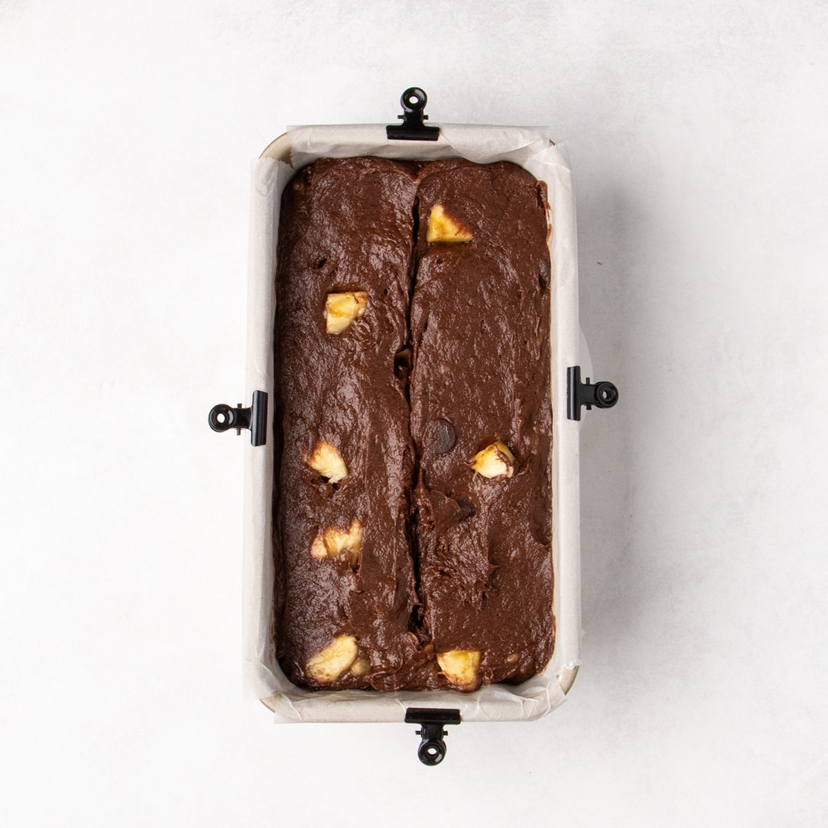 Chocolate batter with visible banana pieces fills a parchment-lined loaf pan secured with black clips.