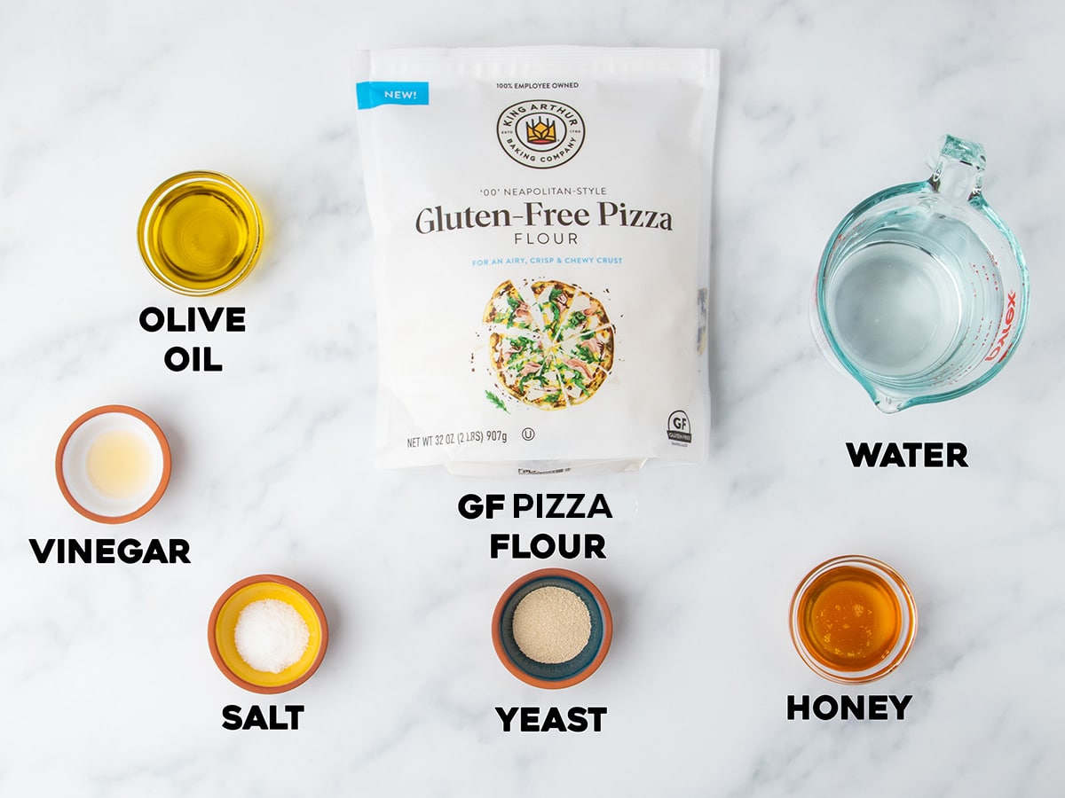Labeled ingredients displayed on a white surface, including bag of gluten free flour, olive oil, honey, yeast, water, vinegar, and salt.