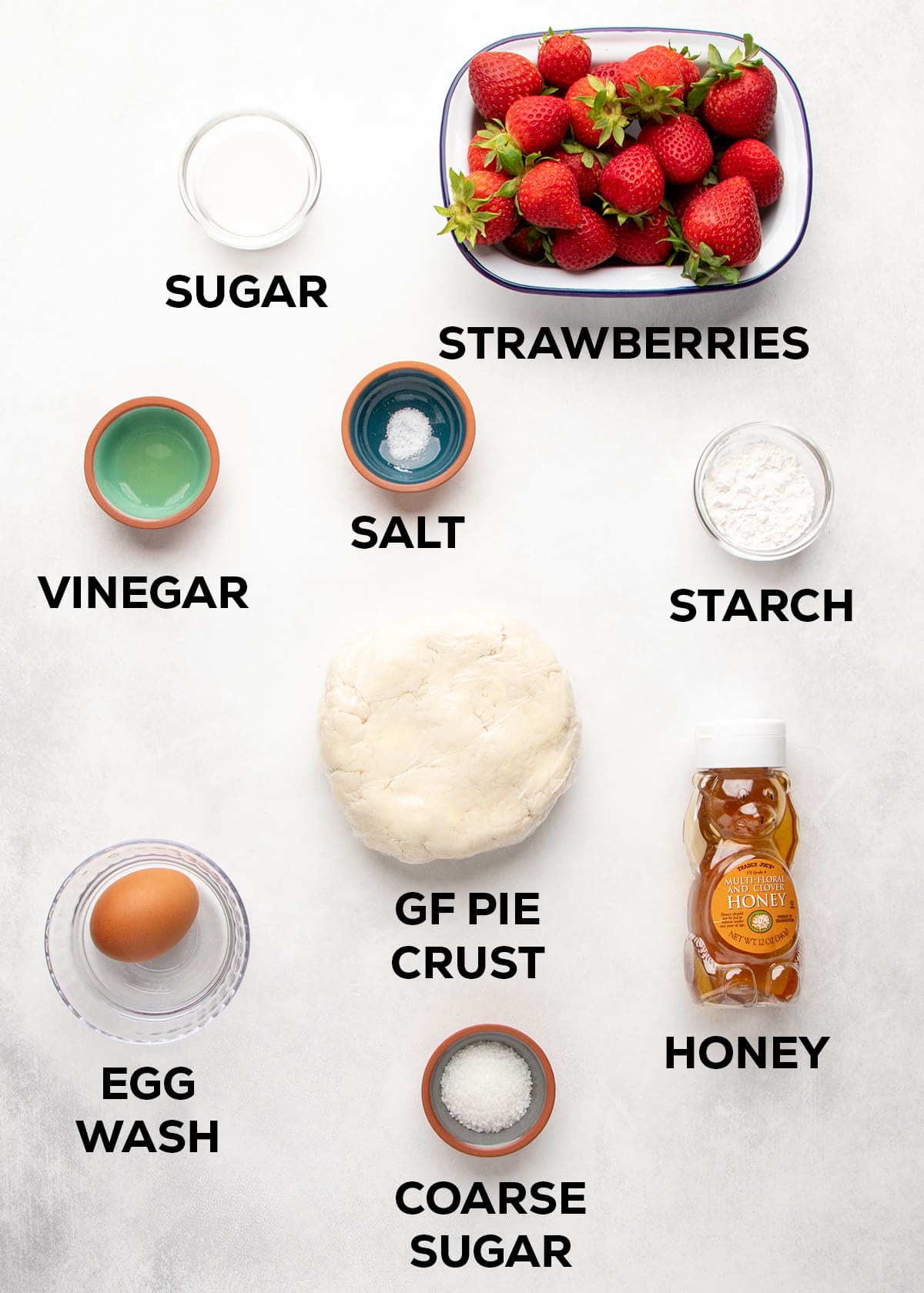 Ingredients labeled sugar, strawberries, vinegar, salt, starch, a disk of gluten free pie crust, a brown egg in a bowl labeled egg wash, honey, and coarse sugar sit on a light surface.