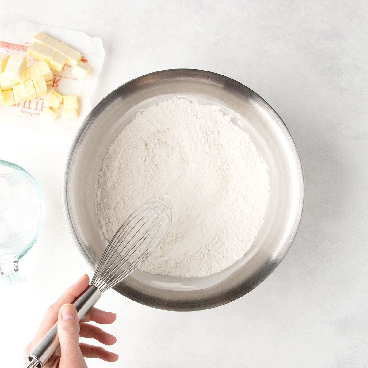 A metal whisk rests in a bowl of white flour mixture, with cubed butter on its wrapper nearby.
