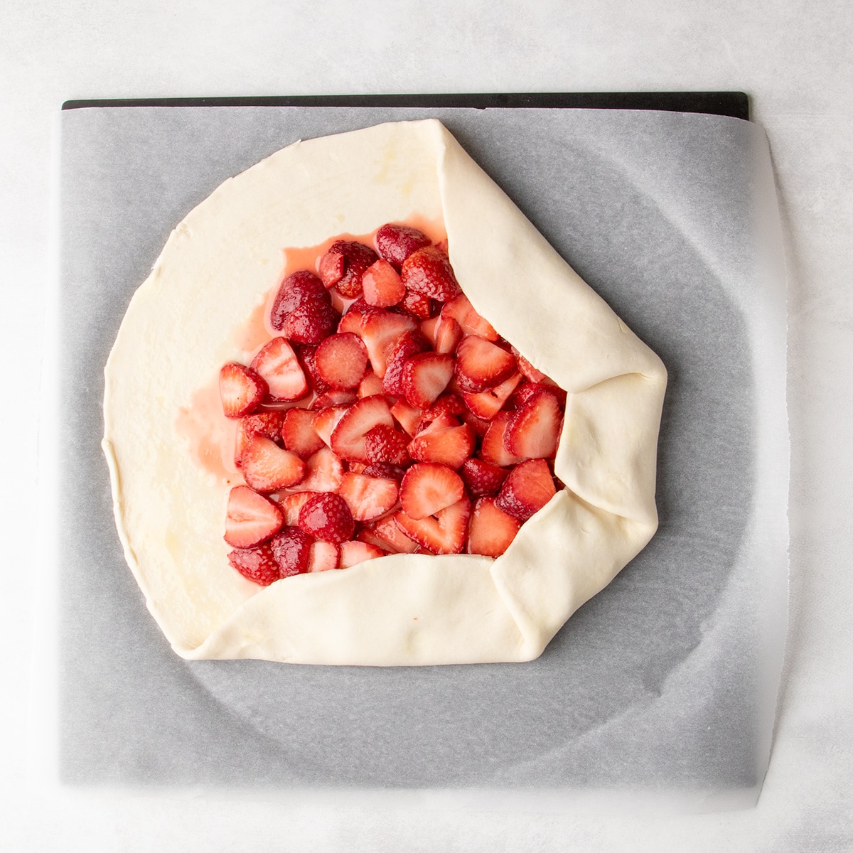 The edges of a round sheet of dough beginning to be folded over sliced strawberries on parchment paper.