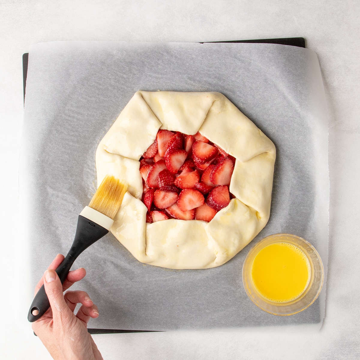 A hand brushes egg wash over the folded edges of an unbaked strawberry galette on parchment paper.