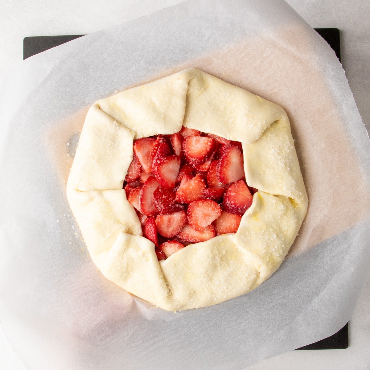 Coarse sugar covers the center and brushed edges of an unbaked strawberry galette on parchment paper on a small brown cutting board underneath.