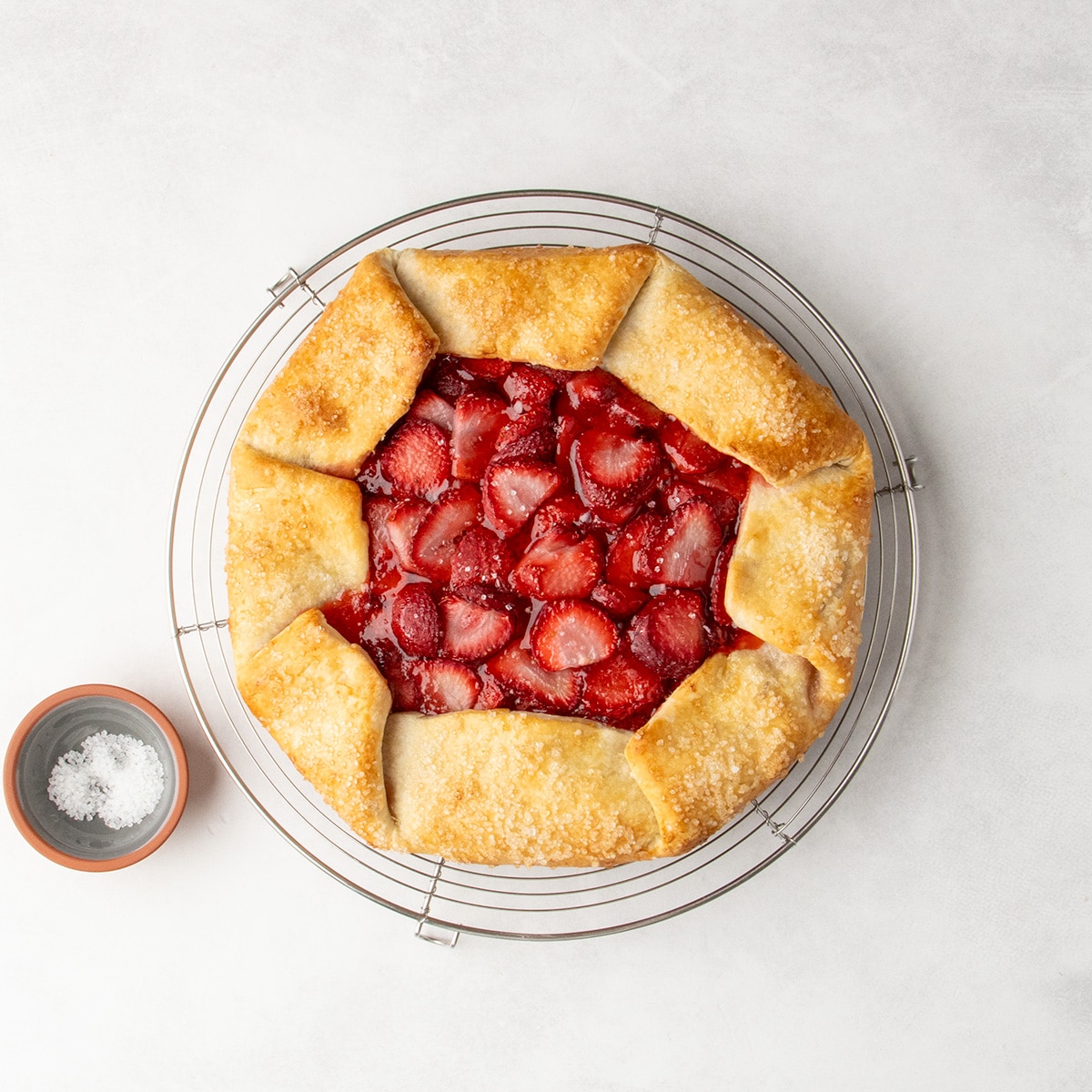 A baked galette with golden brown edges and a jammy sliced strawberry center cools on a round wire rack with a small bowl of coarse sugar nearby.