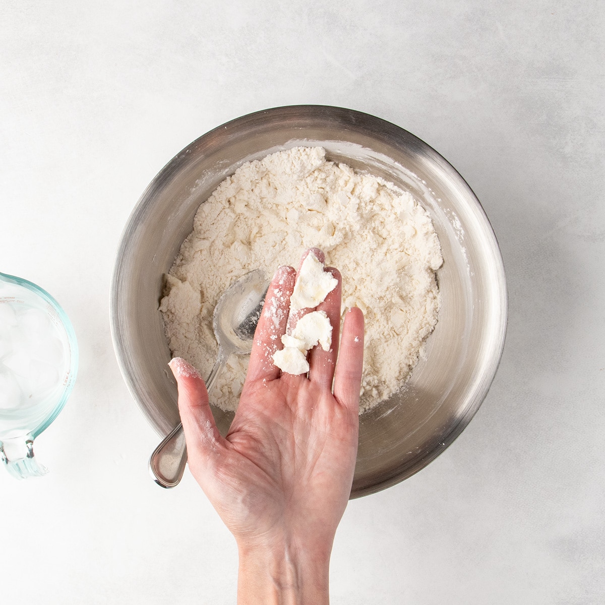 The open palm of a left hand holds pieces of flattened, floury butter over a bowl of flour mixture with a spoon inside.