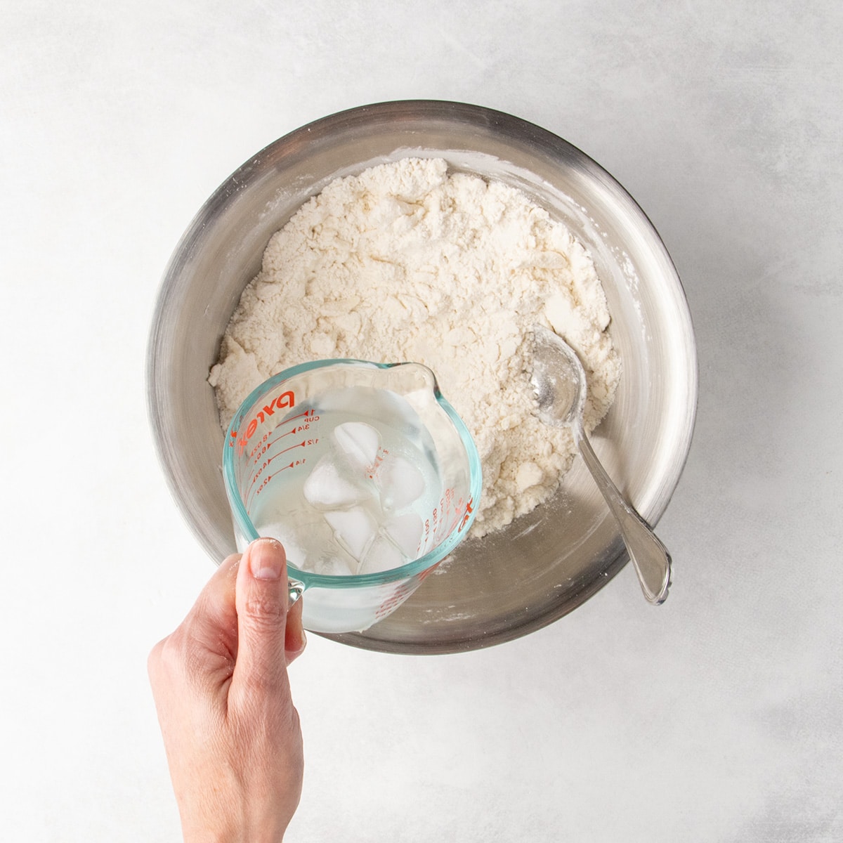 A hand pours ice water from a glass measuring cup into a bowl of flour mixture and butter pieces.