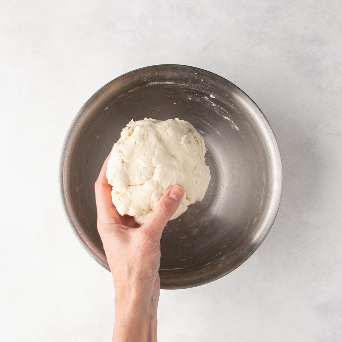 A ball of pale pie crust dough in a hand over a metal mixing bowl.