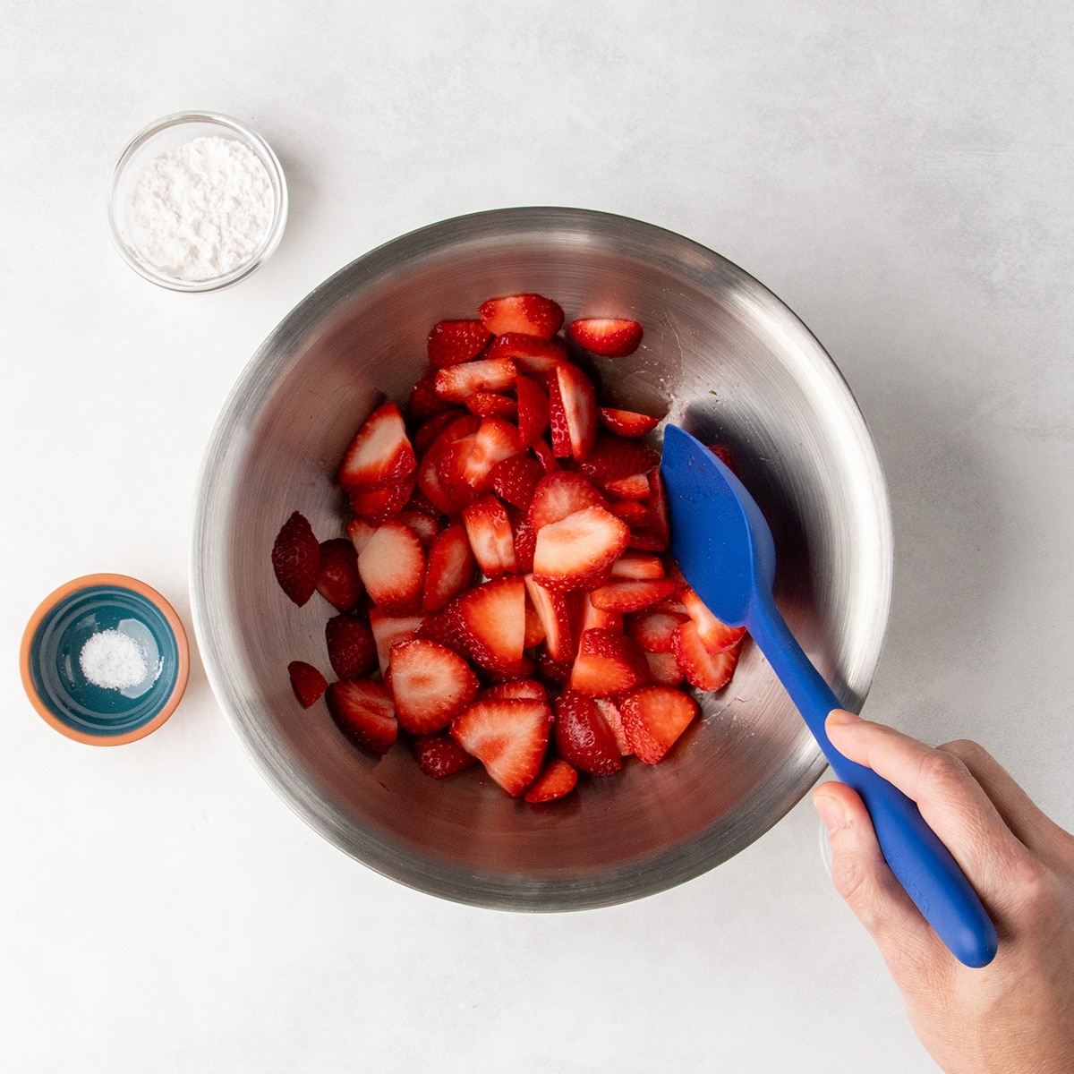 Sliced fresh strawberries sit in a metal mixing bowl with a hand holding a blue spatula resting inside.