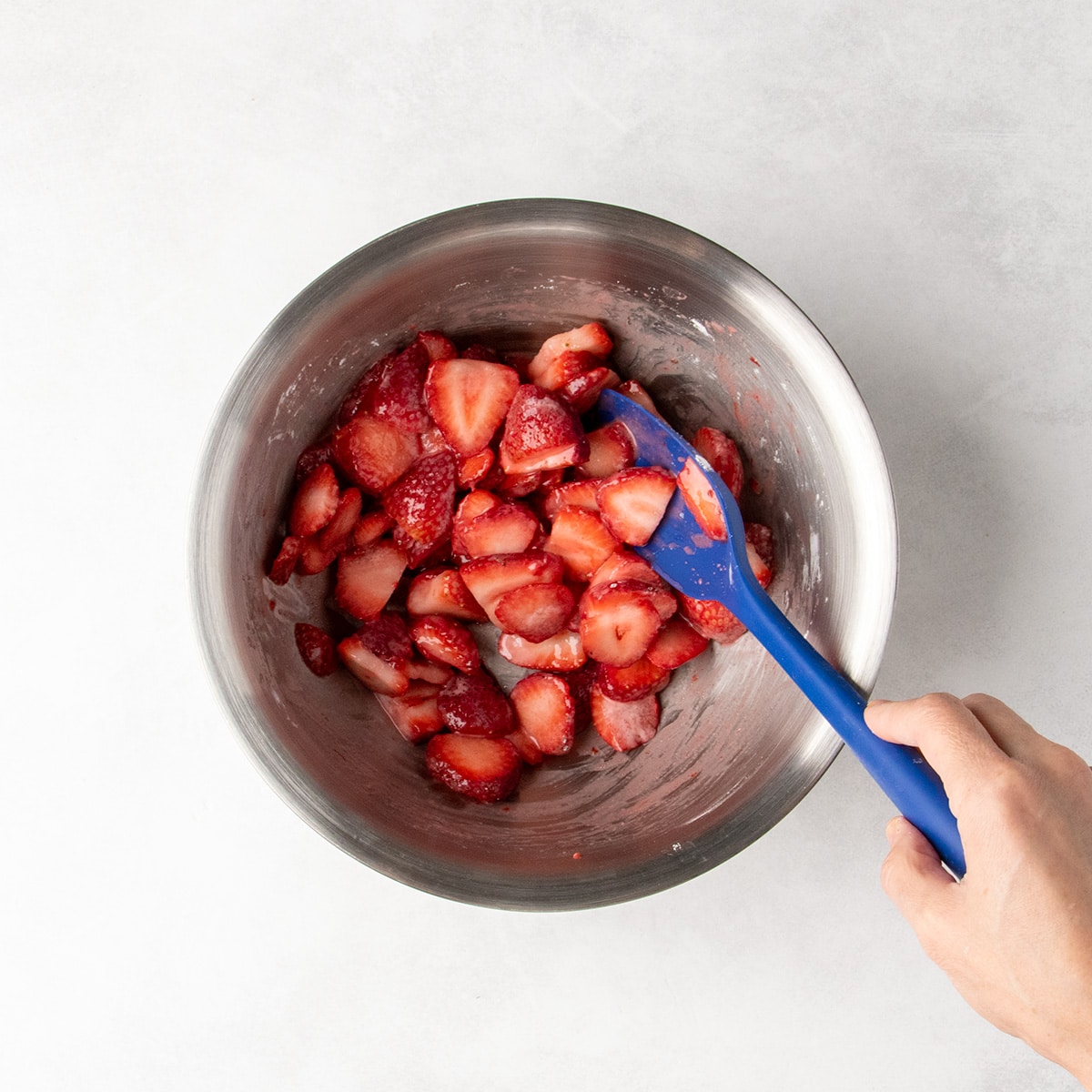 Sliced strawberries in a metal bowl being mixed with a glossy coating with a hand holding a blue spatula.