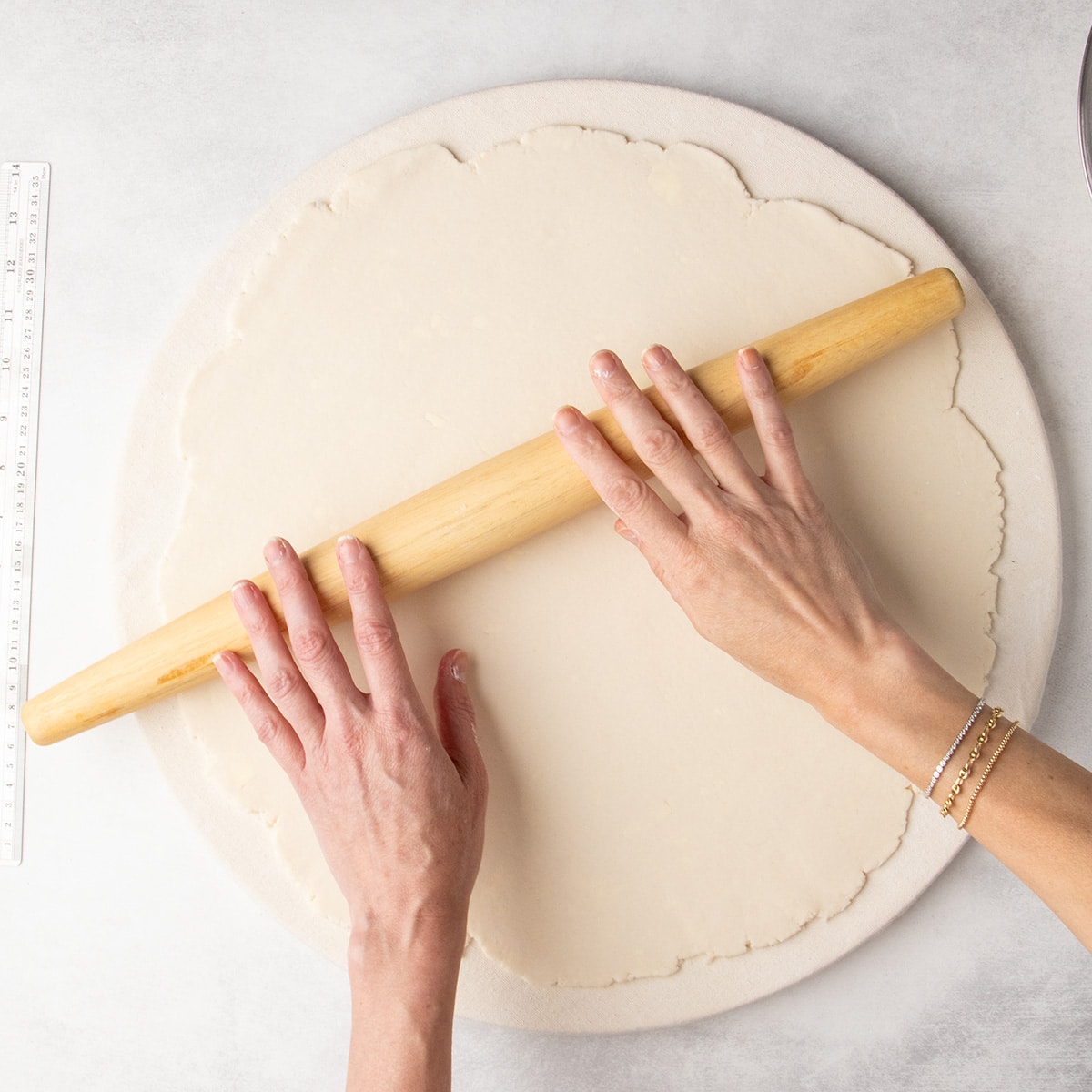 Two hands rolling pale dough into a large round using a wooden tapered rolling pin on a round cloth surface.