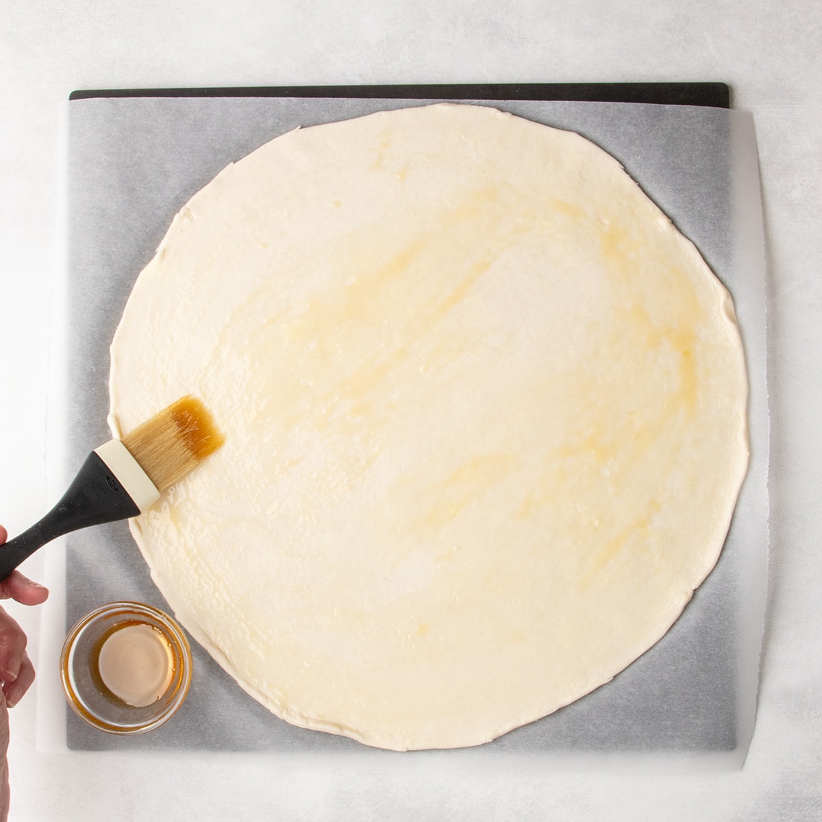 A hand holding a pastry brush, using it to spread light yellow liquid over a round sheet of dough on parchment paper.