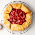 A baked gluten free galette with glossy strawberry filling and golden folded edges sits on a round wire rack with a small bowl of coarse sugar nearby, all on a white surface.