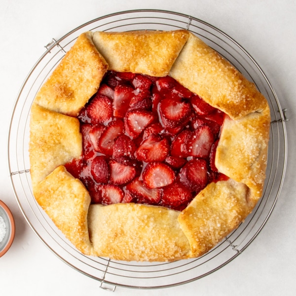A baked gluten free galette with glossy strawberry filling and golden folded edges sits on a round wire rack with a small bowl of coarse sugar nearby, all on a white surface.