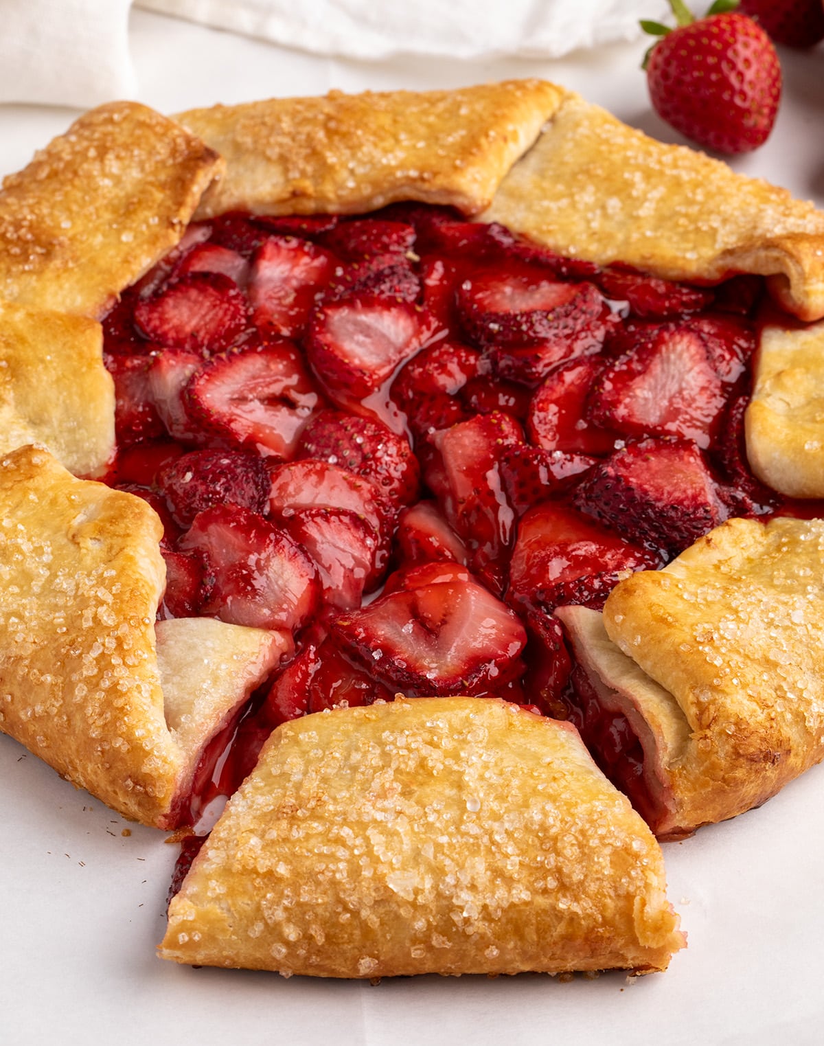 A close view of a baked strawberry galette shows glossy strawberry filling and golden folded pastry edges with coarse sugar and whole strawberries in the background.