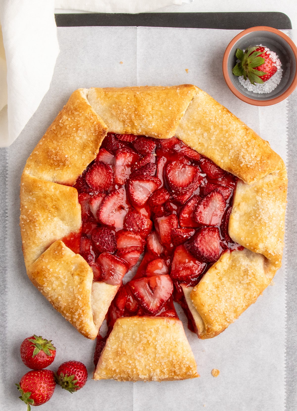 Overhead view of a partially sliced, fully baked strawberry galette sits on parchment paper with whole strawberries and a small bowl nearby.