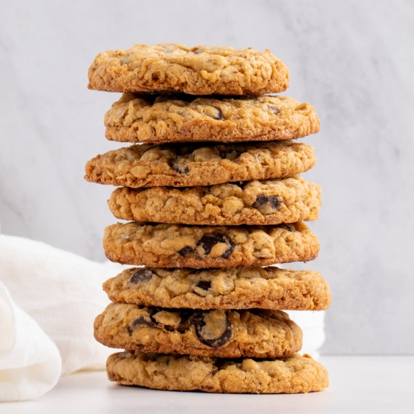 Tall stack of 8 golden brown thick gluten free oatmeal cookies with chocolate chips on light surface with white cloth in background