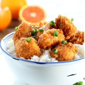 closeup image of glistening, saucy breaded gluten free orange chicken pieces on white rice in a while bowl with chopped green onions