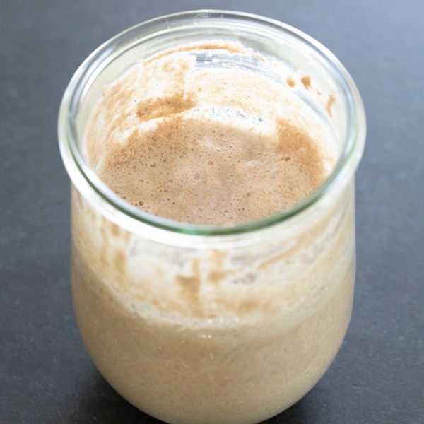 closeup image of light brown gluten free sourdough starter thick liquid light brown in jar on black surface